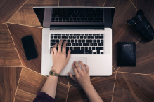 Someone's hands typing on a silver laptop with a black keyboard on a wooden table with a cell phone, camera and wallet surrounding the laptop