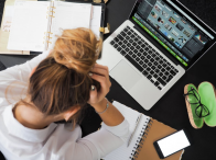 A blonde woman with her hair in a bun looking down in distress with an open laptop, notebooks and other supplies on a desk with her hand on her forehead