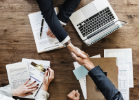 Two people shaking hands over brown table, there is a woman taking notes and there are several other documents on the table. It seems like they are closing a deal and there is also a laptop on the table.