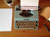 Picture of a teal typewriter on a wooden table with a cup of coffee, pens and stationary by it.