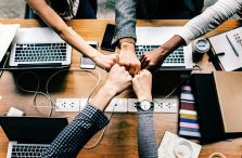 A photo of five hands fist bumping each other over a table of binders, notebooks, smartphones, and laptops.